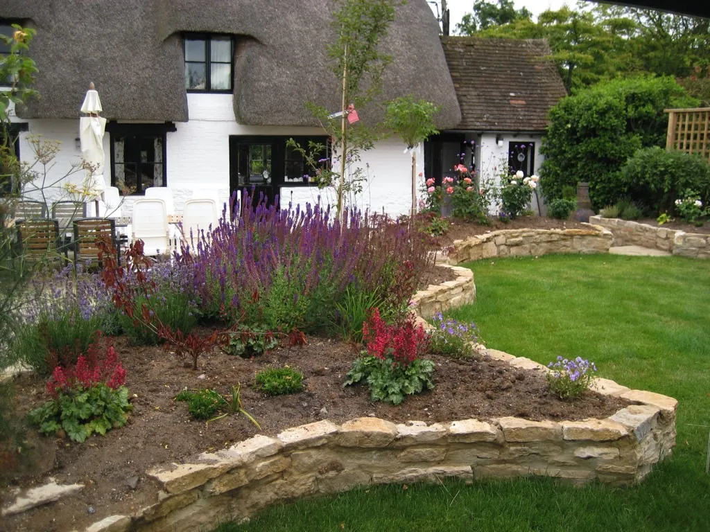 A thatched cottage behind a curved, stone-walled bed of purple and red flowers